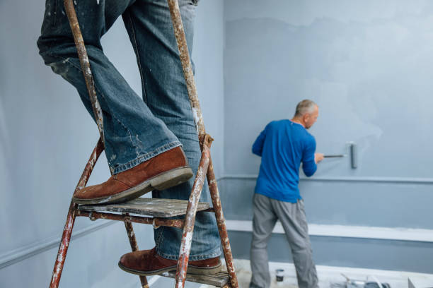 Two male builders are painting a room. One man is using a ladder and only his legs are visible. The other man is in the background.